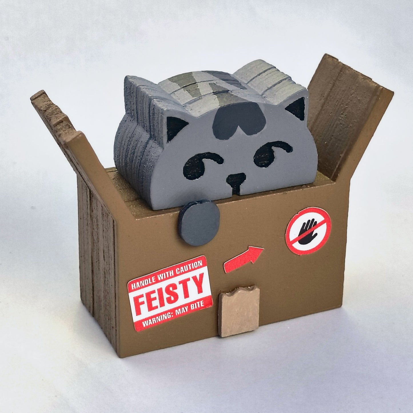 Wood figurine of a gray cat in a cardboard box with shipping label stickers that say "handle with caution, fiesty, warning: may bite" and a do not touch symbol